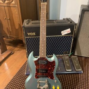 Front view of a customized Epiphone SG Special electric guitar in a metallic blue-grey finish. It features a dark tortoise pickguard, dual humbuckers, yellow control knobs, an aftermarket tremolo system, and a red devil emblem on the headstock. The guitar is displayed in front of a black and blue Vox Valvetronix amplifier.