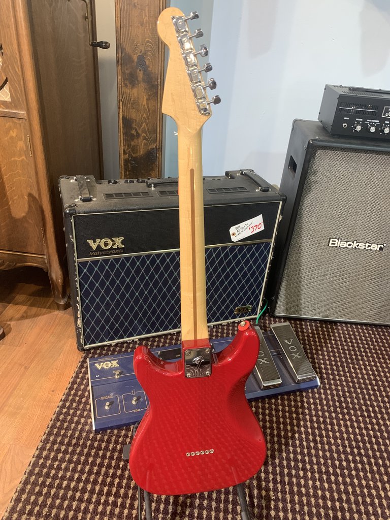 Back view of a red Fender Lead II electric guitar. The photo clearly shows the maple neck, the dark 'skunk stripe' on the back, the chrome six-bolt neck plate, and the string-through-body ferrules. Guitar amplifiers and effects pedals are visible in the background.