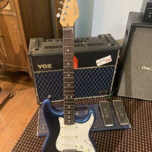 Full front view of a dark blue Fender Stratocaster electric guitar with a white pickguard on a stand, with a Vox Valvetronix amplifier behind it.