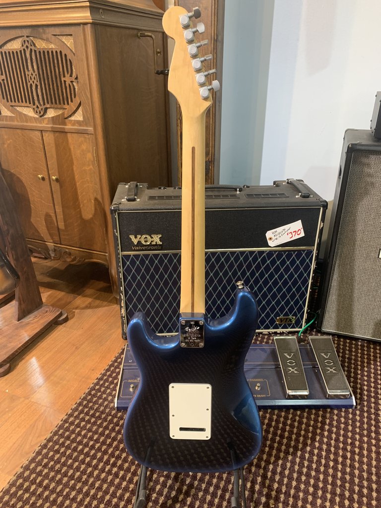 Full rear view of a dark blue Fender Stratocaster electric guitar, showing the maple neck and headstock with chrome tuning machines, with a Vox amplifier behind it.